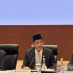 Seorang pejabat berseragam jas dan peci sedang berbicara di meja panel rapat, dengan dua orang lain di sampingnya