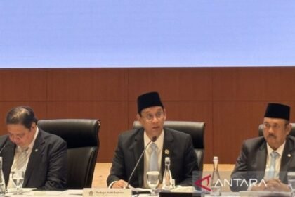 Seorang pejabat berseragam jas dan peci sedang berbicara di meja panel rapat, dengan dua orang lain di sampingnya