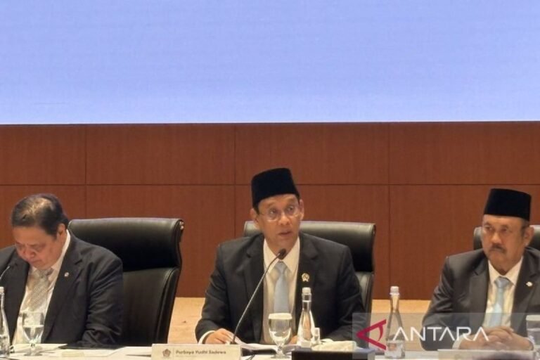 Seorang pejabat berseragam jas dan peci sedang berbicara di meja panel rapat, dengan dua orang lain di sampingnya