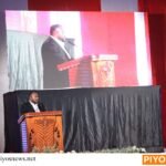 Seorang pembicara berdiri di podium kayu dengan layar besar menampilkan wajahnya di belakang, bendera Indonesia terlihat di samping.