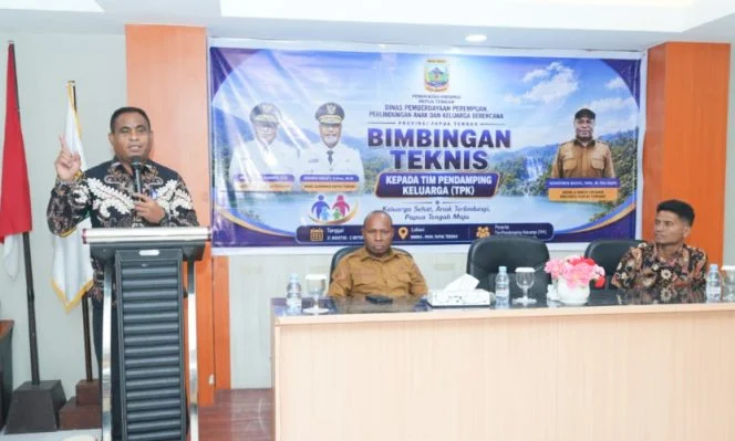 Pembicara berdiri di podium di depan spanduk Bimbingan Teknis Tim Pendamping Keluarga, tiga orang duduk di meja depan