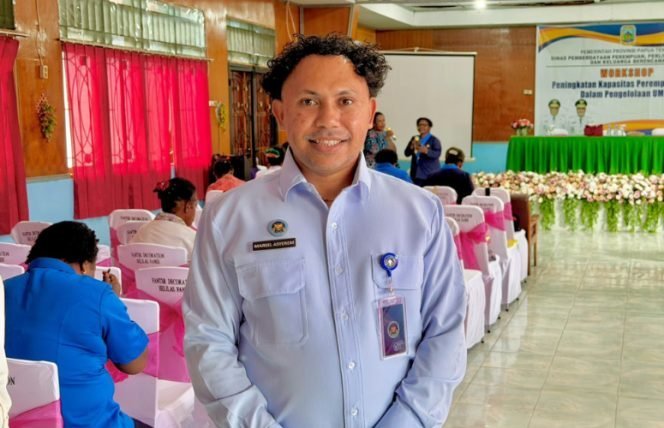 Marsel Asyerem berkemeja biru muda berdiri di aula pelatihan dengan spanduk workshop di belakangnya